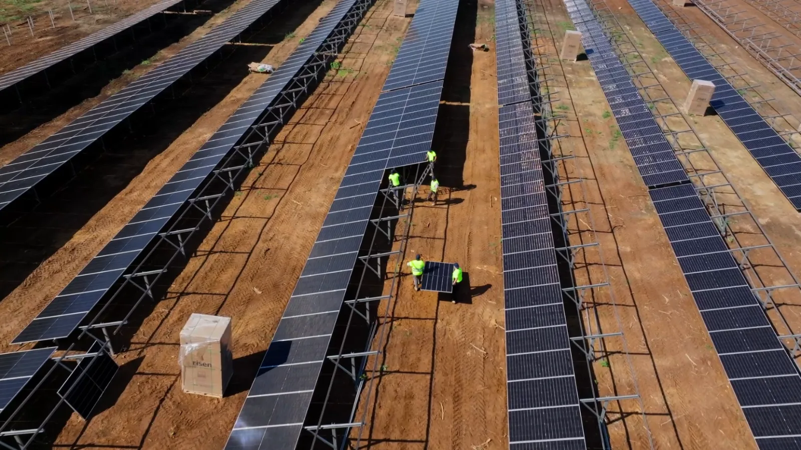 Vista aérea de operarios con chaleco amarelo montando paneis solares sobre as estruturas da planta, en terra vermella.