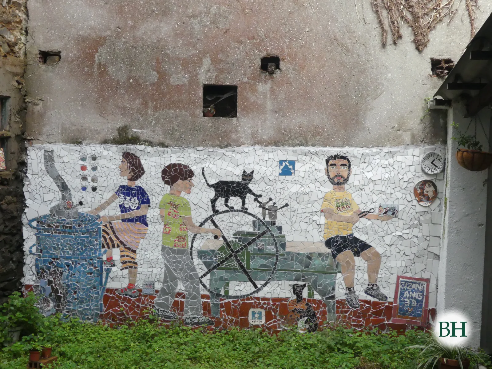 Mosaico en la pared del patio del estudio, sobre la hierba: una mujer junto a un horno azul, un hombre que mueve la rueda de un tórculo con un gato negro encima, otro gato sentado debajo y un hombre con barba que hace mosaico.