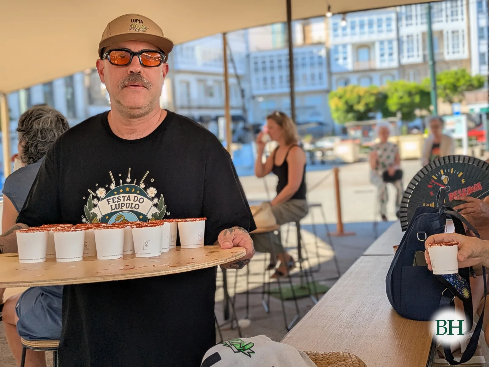 Un home coa camiseta da Festa do Lúpulo leva unha bandexa con vasos de degustación baixo a carpa