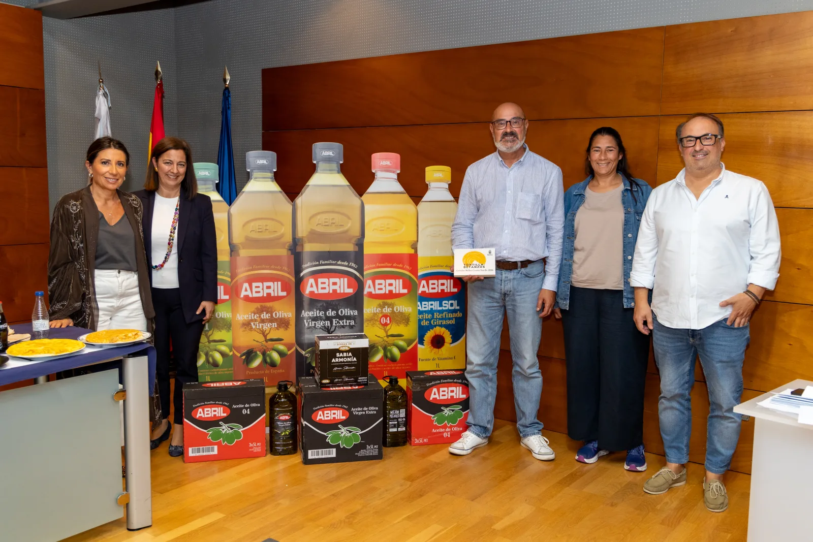 Seis personas de pie junto a botellas gigantes y cajas de Aceites Abril. A la izquierda, una mesa con dos tortillas en platos.