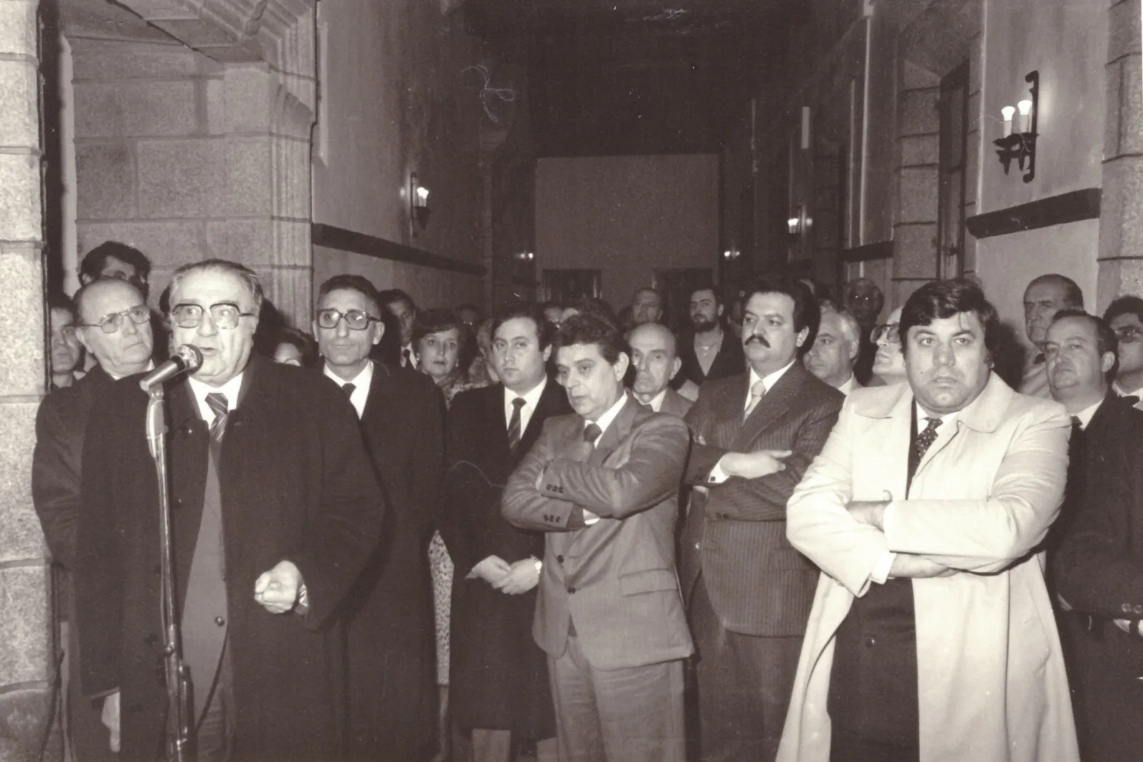 Fotografía en branco e negro dun numeroso grupo de persoas nun corredor de pedra escoitando un home que fala diante dun micrófono