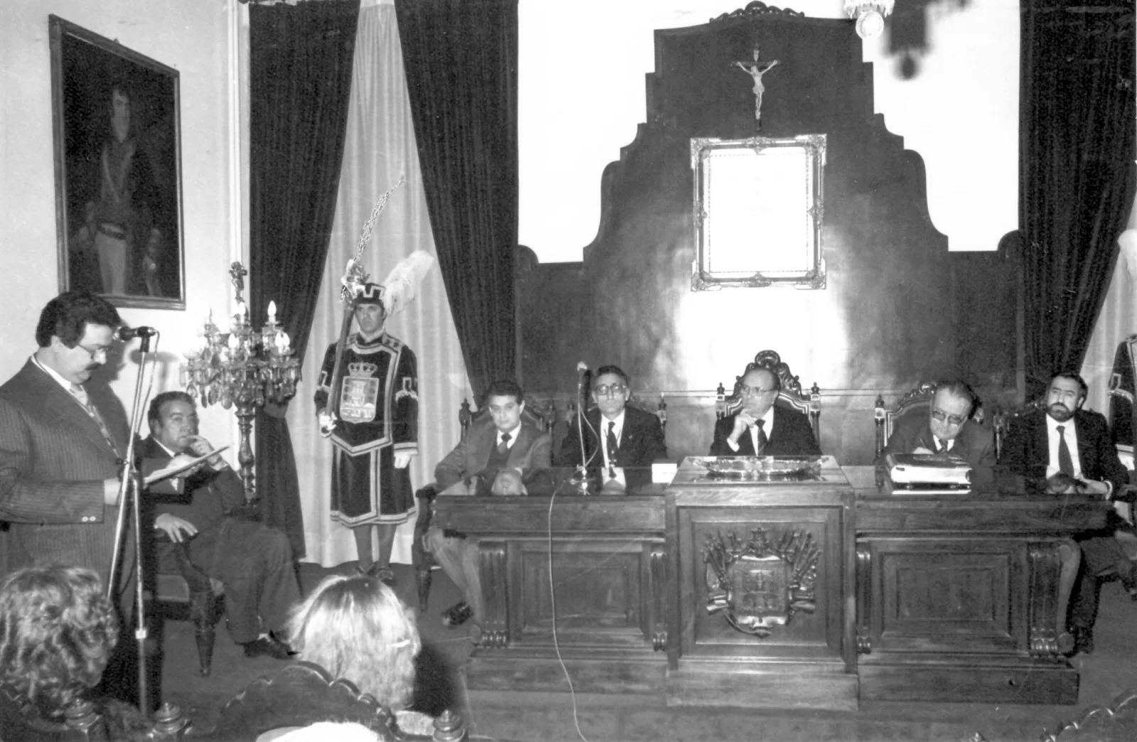 Fotografía en branco e negro dun acto na Sala Capitular do Concello de Betanzos: un home le diante dun micrófono mentres as autoridades ocupan a mesa presidencial