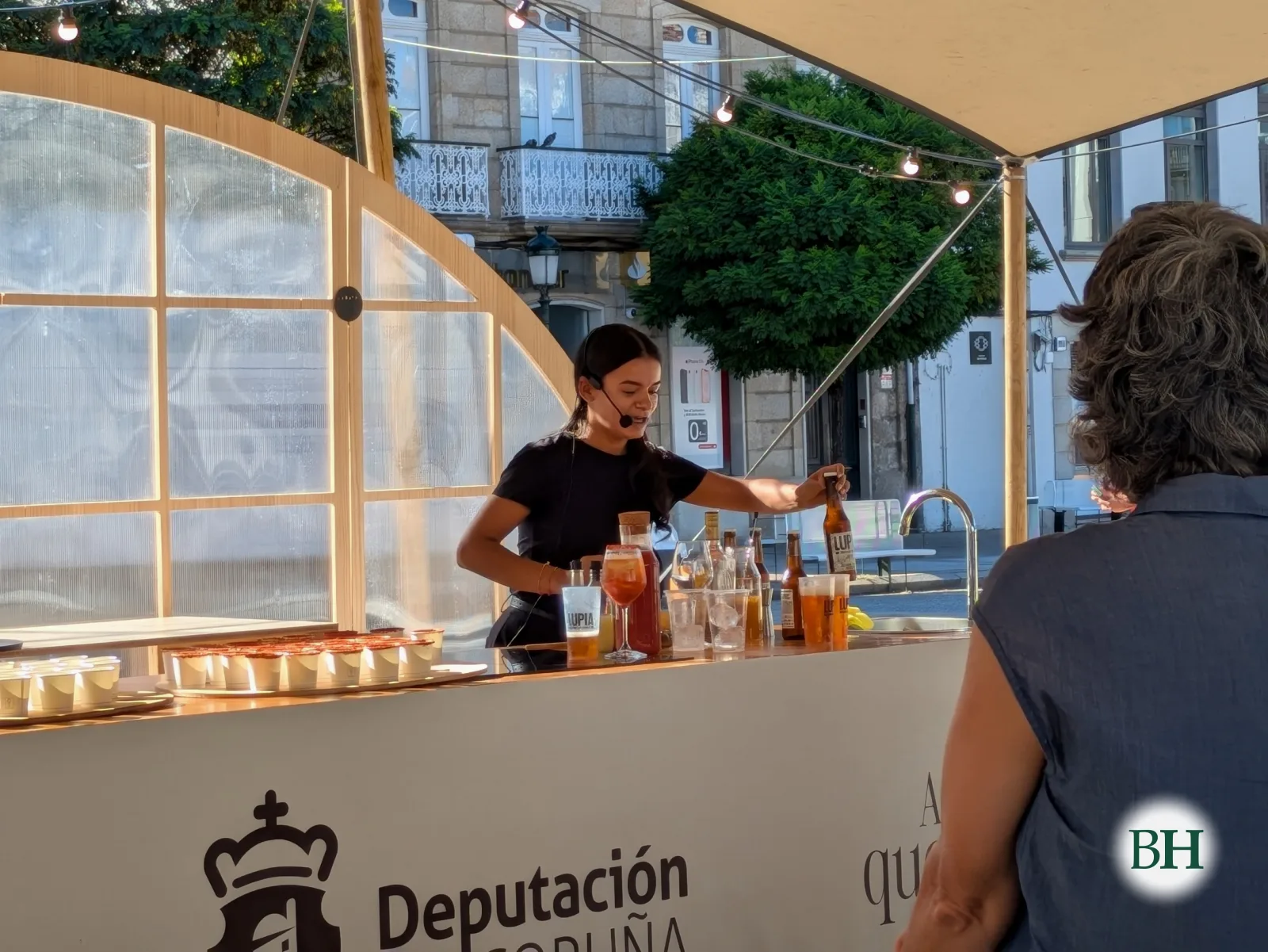 Unha cociñeira serve unha copa nun showcooking na barra da carpa da Deputación da Coruña, con botellas de Lupia diante