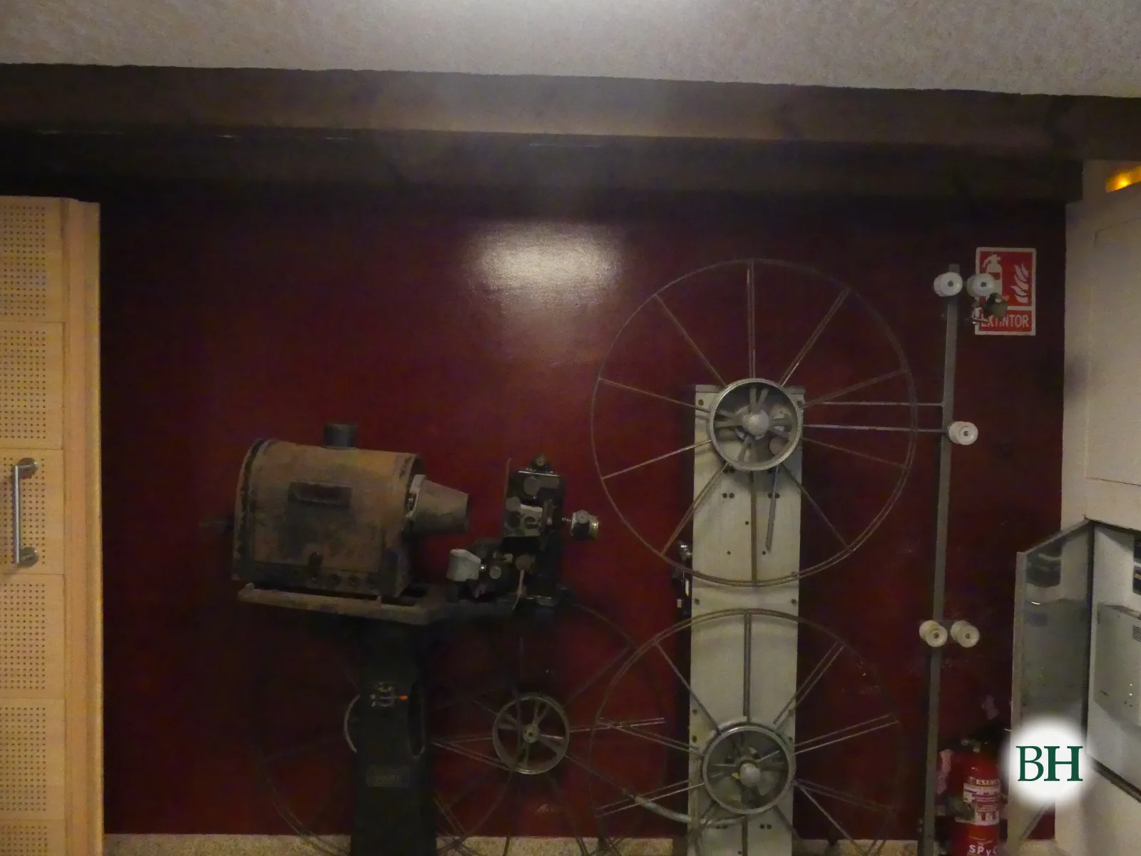 Un proxector cinematográfico antigo e dúas grandes bobinas expostos contra unha parede granate.