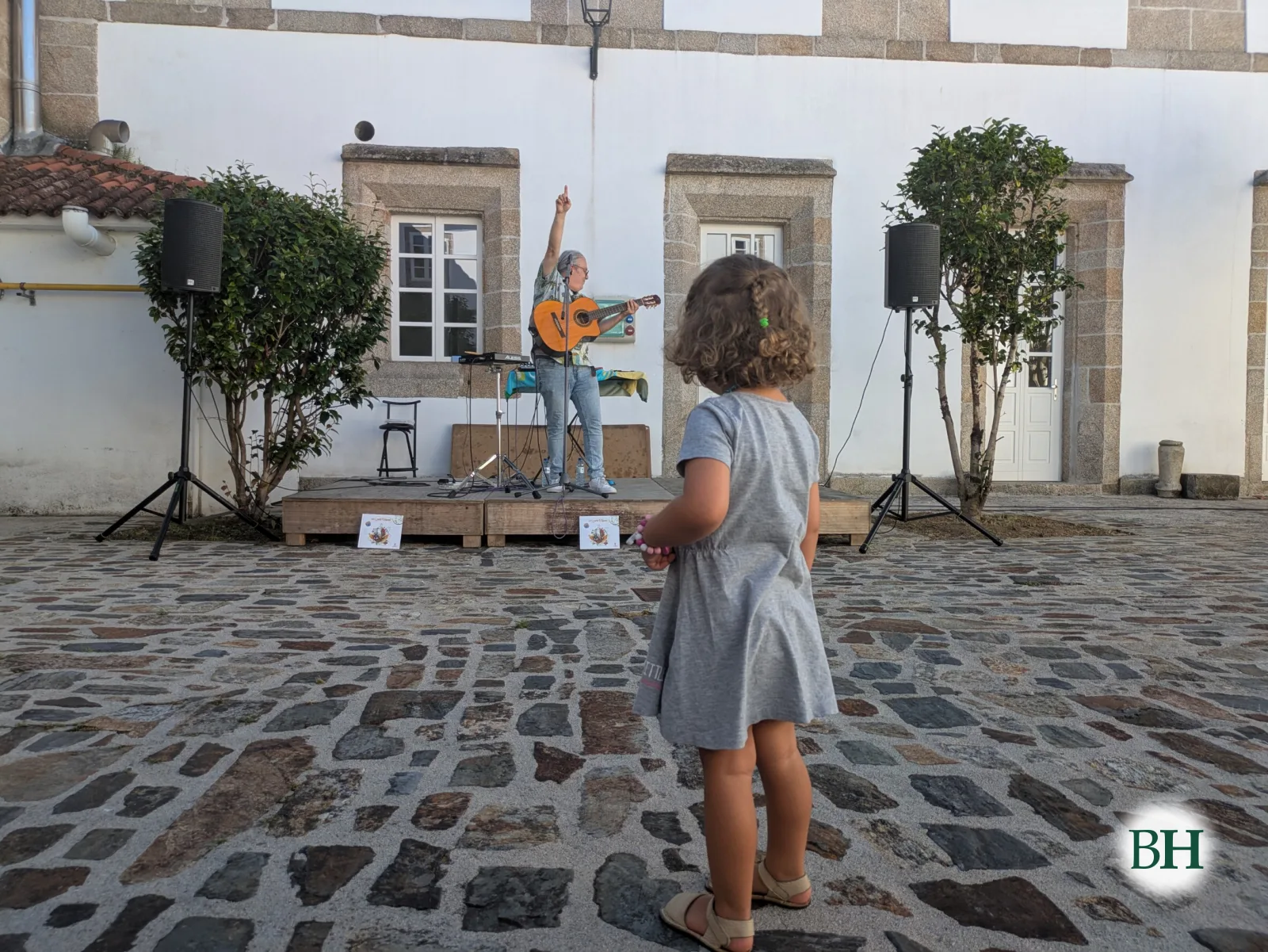 Unha nena, de costas, mira a Chuches Amil, que toca a guitarra co brazo en alto sobre o escenario montado no patio do Museo das Mariñas