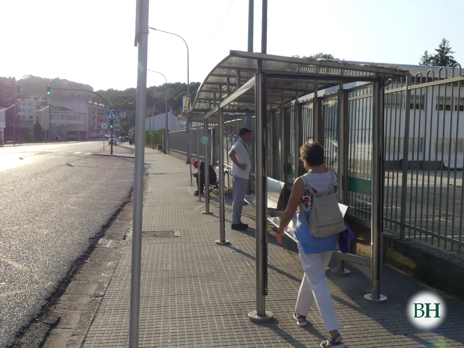 Parada de autobús con marquesiña metálica, dúas persoas camiñando pola beirarrúa e unha rúa con semáforo e edificios ao fondo con luz de solpor.