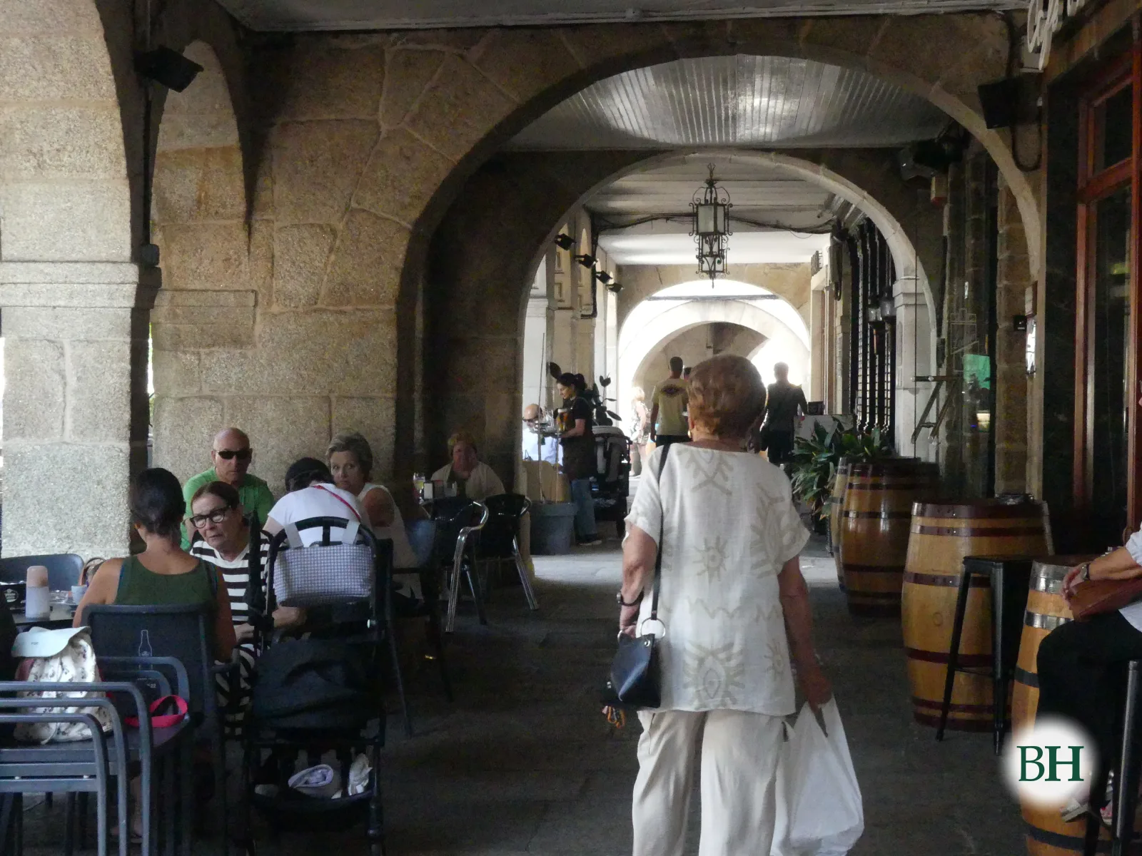 Galería de pedra con sucesión de arcos, terraza de bar con mesas, cadeiras e barrís de madeira, e varias persoas sentadas ou camiñando.