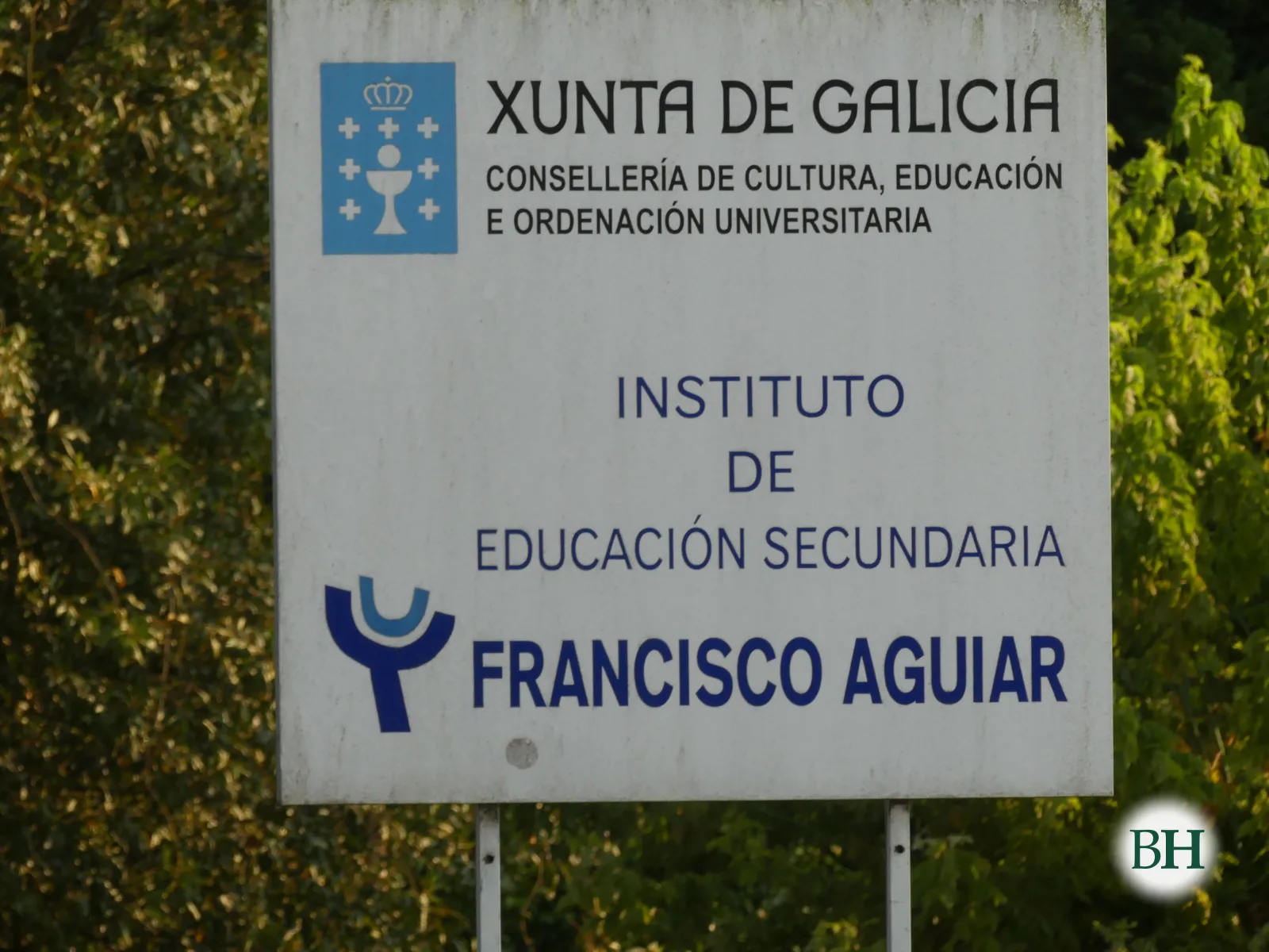 Cartel institucional blanco con escudo y el texto Xunta de Galicia, Consellería de Cultura, Educación e Ordenación Universitaria, Instituto de Educación Secundaria Francisco Aguiar, entre vegetación.