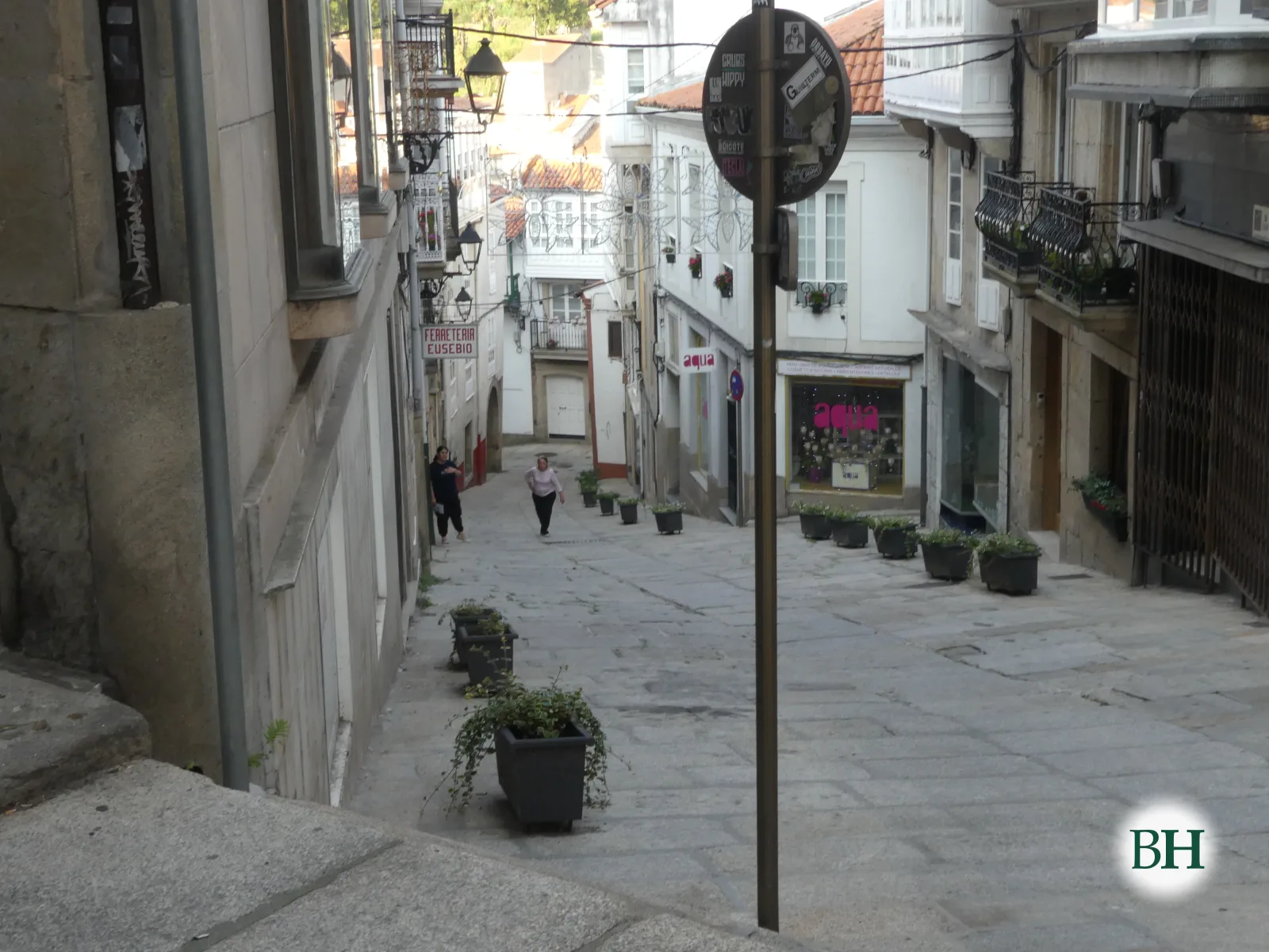 Calle peatonal en pendiente con macetas de plantas, farola metálica y el rótulo de una ferretería, con dos personas caminando.