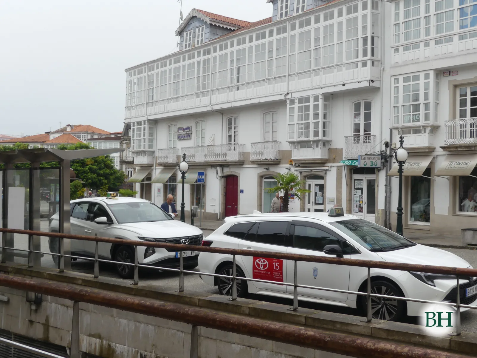 Parada de taxis con dos coches blancos frente a un edificio de galerías acristaladas y una palmera.