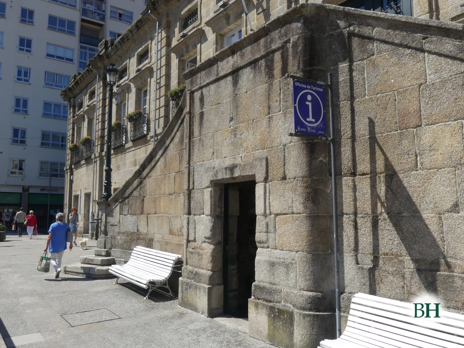 Fachada de pedra labrada con porta en arco e cartel azul de oficina de turismo, farola negra, bancos brancos e peóns xunto ao edificio.