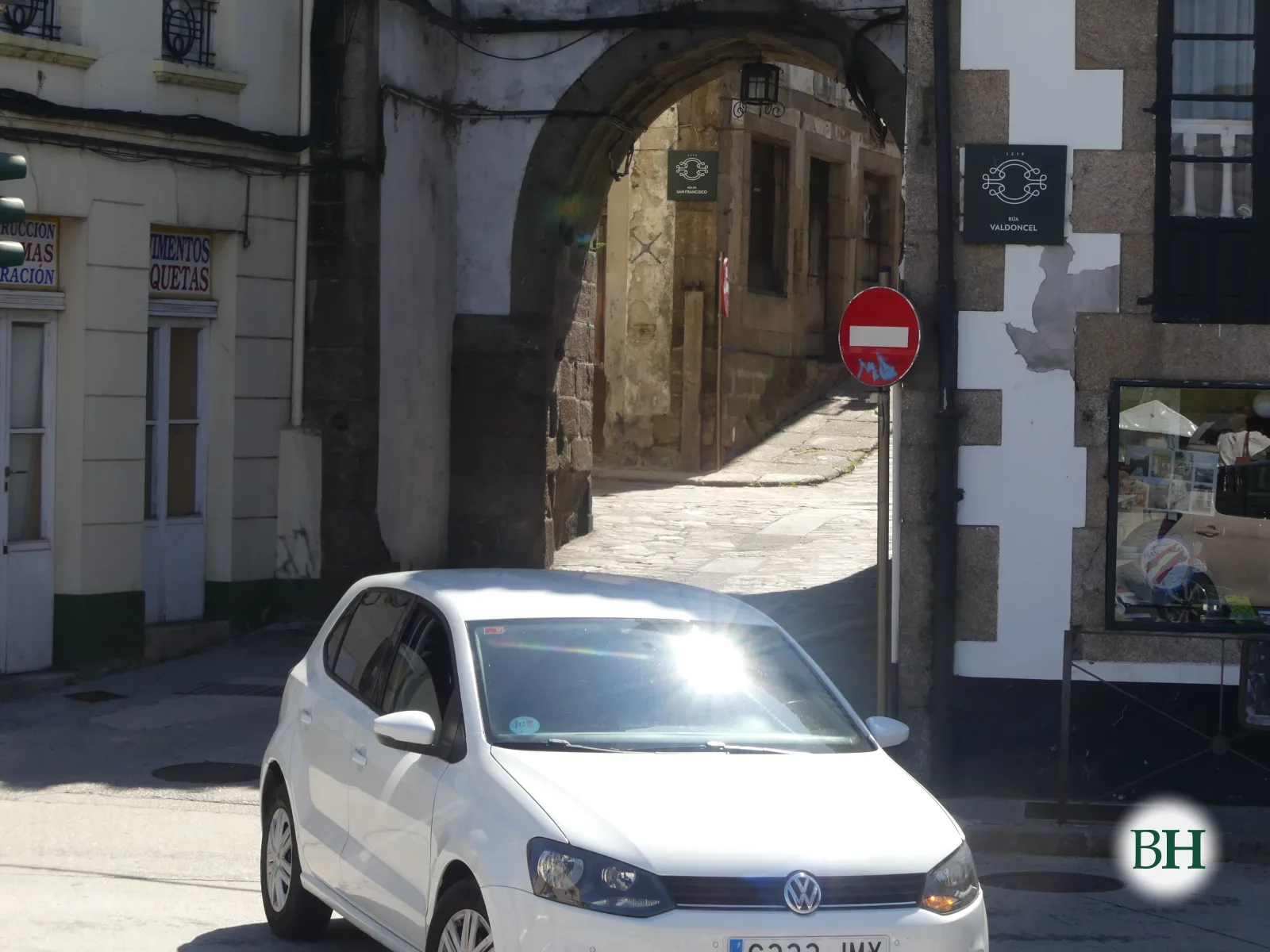 Automóbil branco aparcado fronte a un arco de pedra que dá acceso a unha rúa empedrada, con fachadas antigas e un sinal de dirección prohibida.