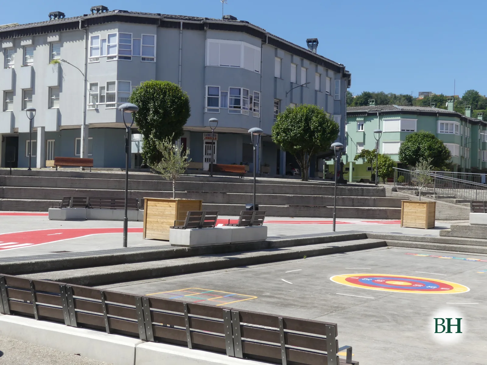 Praza urbana moderna con pavimento pintado, bancos de madeira, xardineiras con árbores e edificios residenciais arredor.