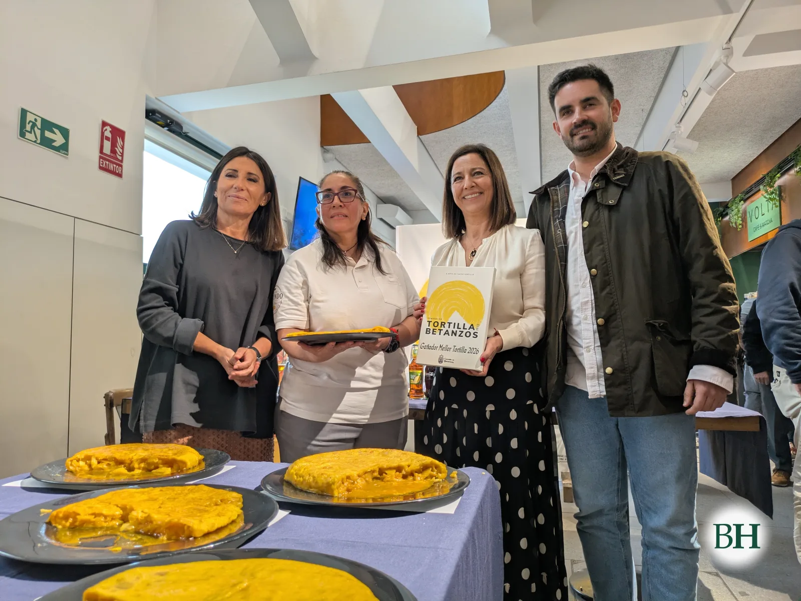 Sheila Delgado, del Mesón Café de Segundo, con polo blanco y un plato de tortilla, sonriente con otras tres personas detrás de una mesa con mantel azul y cinco tortillas. Una mujer sostiene un cuadro amarillo de Ganador Mejor Tortilla 2026.