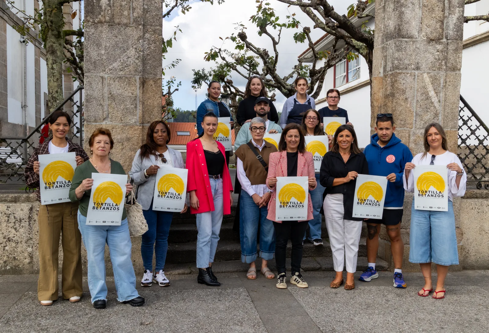 Presentación da XVI Semana da Tortilla de Betanzos: unha quincena de persoas, hostaleiras e hostaleiros participantes e representantes municipais, posan entre dous piares de pedra sostendo o cartel amarelo do evento.
