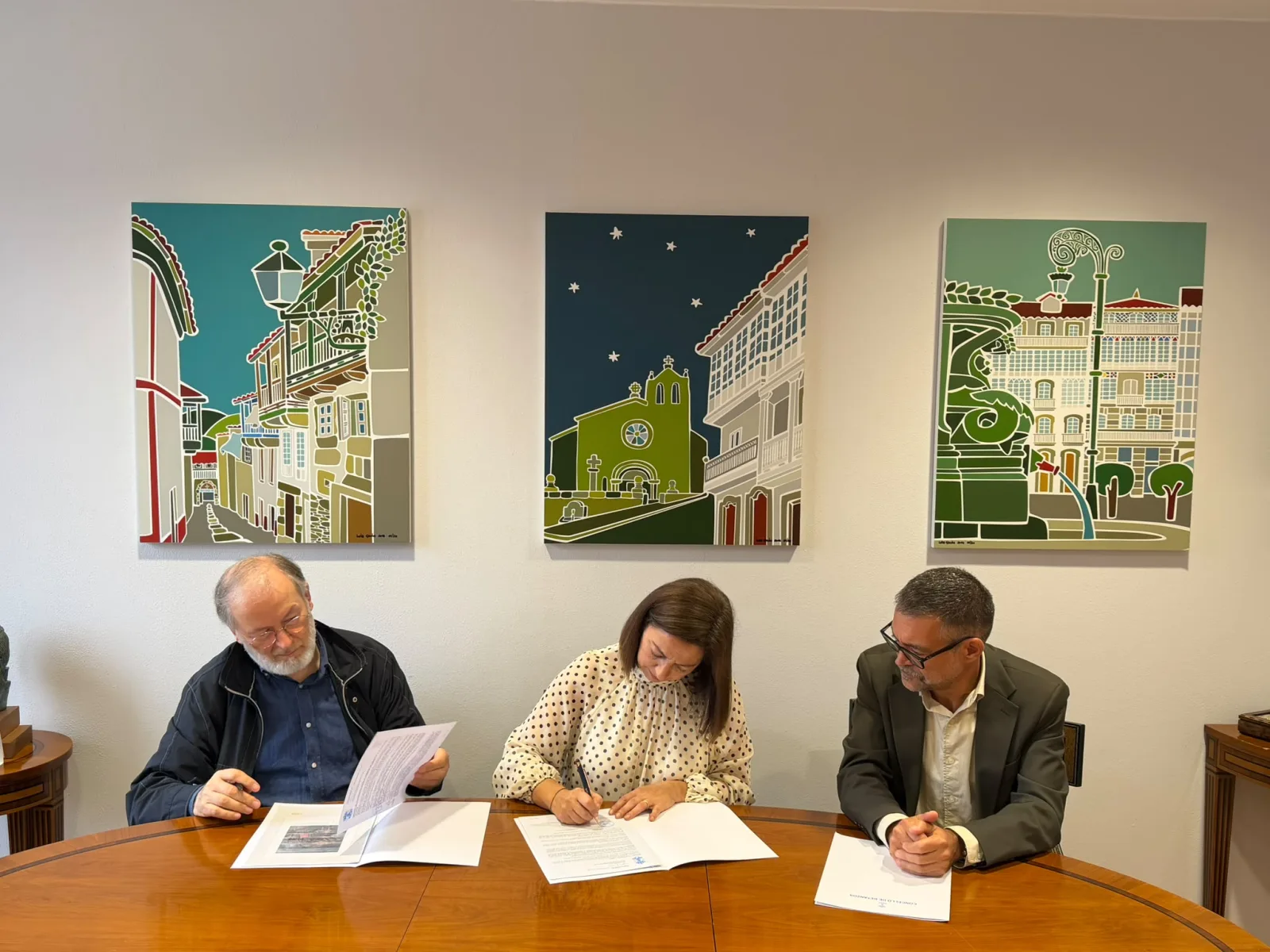 A alcaldesa, María Barral, asina o convenio para restaurar o telón do Alfonsetti xunto ao presidente da Fundación L. Monteagudo, Alfredo Erias, que le o documento, baixo tres cadros de rúas de Betanzos.