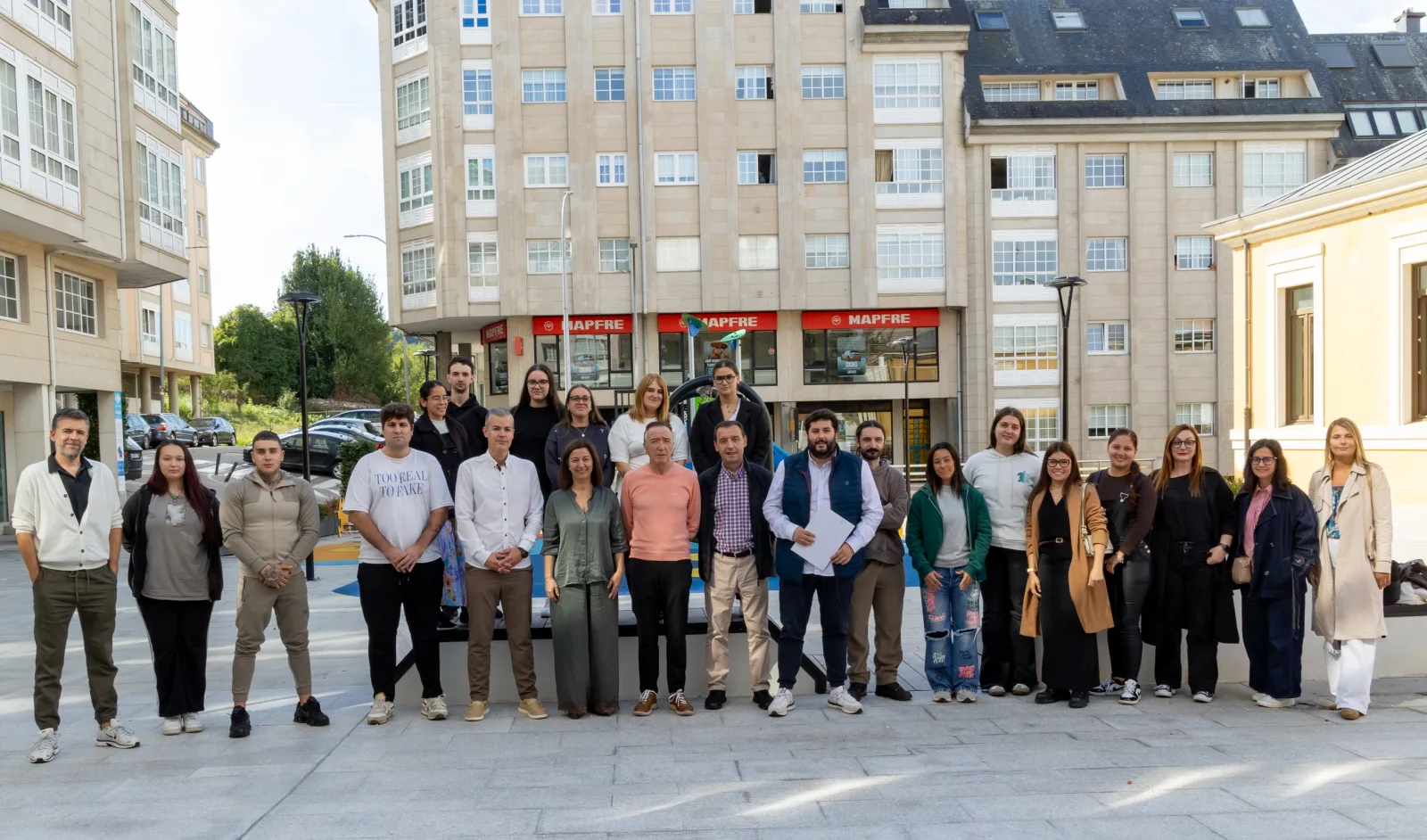 Foto de grupo del alumnado y de los representantes municipales del nuevo obradoiro de emprego sociosanitario, de pie en una plaza de Betanzos con edificios de viviendas al fondo.